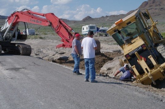 The bucket fills in either side of the trench, while thick cables are fastened to the grader's axle.